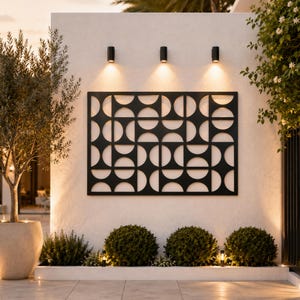 May include: A rectangular black metal wall art panel featuring a repeating geometric pattern of semicircles and circles. The piece is mounted on a white exterior wall, illuminated by three small black cylindrical wall-mounted light fixtures.