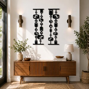 Może przedstawiać: Dwa czarne metalowe elementy dekoracji ściennej w stylu mid-century modern. Każdy element ma pionowy układ geometrycznych kształt&oacute;w, w tym prostokąt&oacute;w, owali i k&oacute;ł. Sztuka jest zamontowana na białej ścianie.