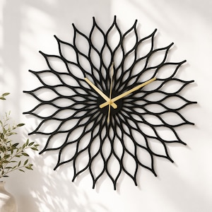 Puede incluir: Un reloj de pared decorativo negro con un diseño floral. El reloj tiene forma circular con intrincados recortes en forma de hoja. Las manecillas del reloj son doradas. El reloj está montado en una pared blanca.