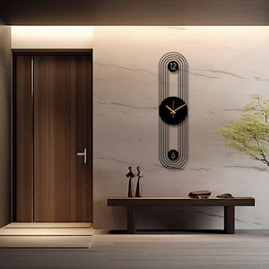May include: A black and gold modern wall clock with a circular face and Roman numeral markings. The clock is hanging on a white marble wall with a wooden door and bench in the background.