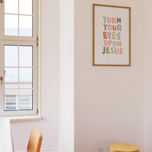 May include: A framed print with the text "TURN YOUR EYES UPON JESUS" in colorful bubble letters. The print is in a room with a window, wooden chairs, and a small wooden stool. The frame is a light brown color.