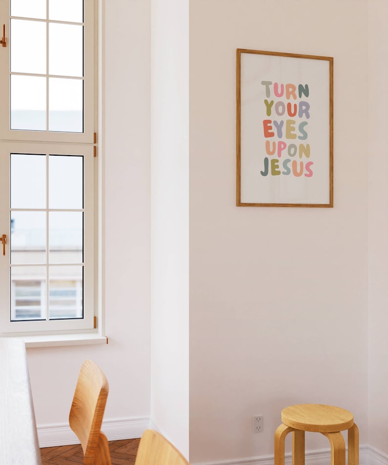 May include: A framed print with the text "TURN YOUR EYES UPON JESUS" in colorful bubble letters. The print is in a room with a window, wooden chairs, and a small wooden stool. The frame is a light brown color.