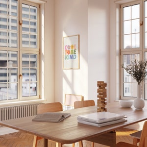 May include: A light wood dining table with a stack of books and a wooden block tower on the surface. Two chairs are visible, one with a book on it. The table is in a room with two large windows and a framed print on the wall that says "Cool to be kind" in colorful letters.