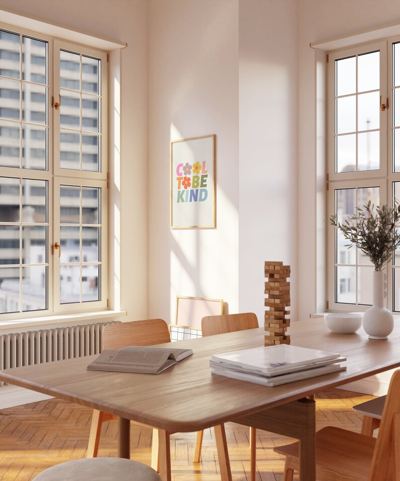 May include: A light wood dining table with a stack of books and a wooden block tower on the surface. Two chairs are visible, one with a book on it. The table is in a room with two large windows and a framed print on the wall that says "Cool to be kind" in colorful letters.
