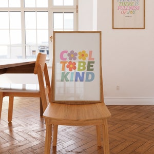 May include: A framed print with the words "Cool to be Kind" in colorful letters with flowers between the words. The print is hanging on a wall in a room with a wooden floor and two chairs.