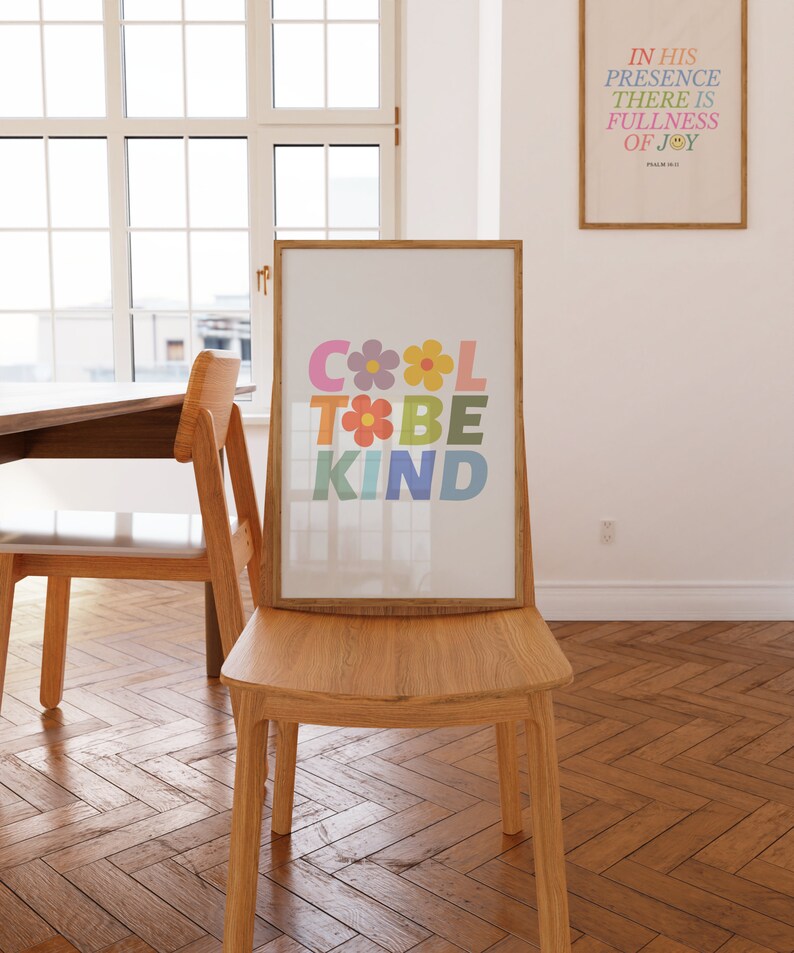 May include: A framed print with the words "Cool to be Kind" in colorful letters with flowers between the words. The print is hanging on a wall in a room with a wooden floor and two chairs.