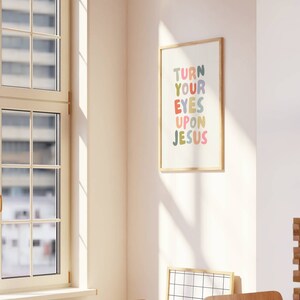 May include: A framed art print with the text "TURN YOUR EYES UPON JESUS" in colorful bubble letters. The print is in a light wood frame and hangs on a white wall. The room has a window and wooden chairs.