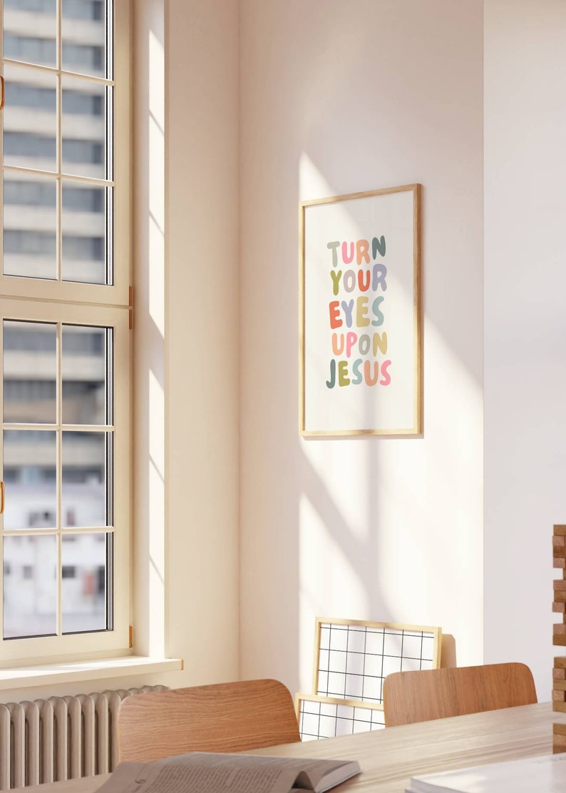 May include: A framed art print with the text "TURN YOUR EYES UPON JESUS" in colorful bubble letters. The print is in a light wood frame and hangs on a white wall. The room has a window and wooden chairs.