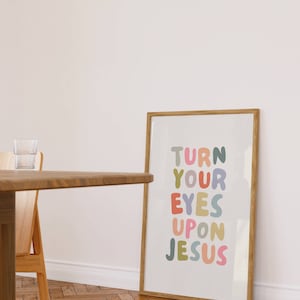 May include: A framed print with the words "TURN YOUR EYES UPON JESUS" in colorful, rounded letters. The print is in a light wooden frame and leans against a white wall. A wooden table and chair are in the foreground.