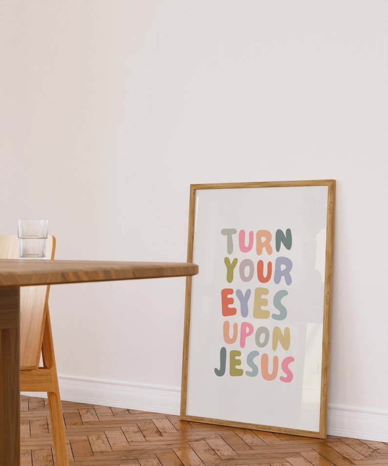 May include: A framed print with the words "TURN YOUR EYES UPON JESUS" in colorful, rounded letters. The print is in a light wooden frame and leans against a white wall. A wooden table and chair are in the foreground.
