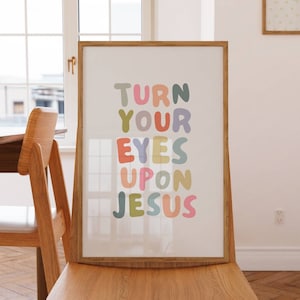 May include: A framed art print with the words "TURN YOUR EYES UPON JESUS" in colorful bubble letters. The print is displayed on a wooden chair, with a window and table in the background. The artwork has a light, airy feel.