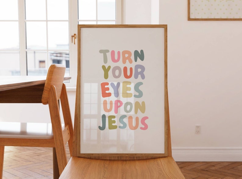May include: A framed art print with the words "TURN YOUR EYES UPON JESUS" in colorful bubble letters. The print is displayed on a wooden chair, with a window and table in the background. The artwork has a light, airy feel.
