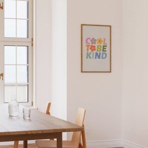 May include: A framed print with the text "COOL TO BE KIND" in a rainbow of colors with flowers in between the letters. The print is hanging on a white wall in a room with a wooden table and chairs.