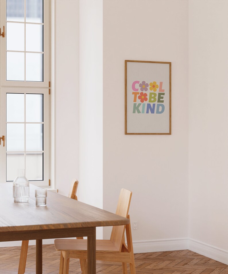 May include: A framed print with the text "COOL TO BE KIND" in a rainbow of colors with flowers in between the letters. The print is hanging on a white wall in a room with a wooden table and chairs.