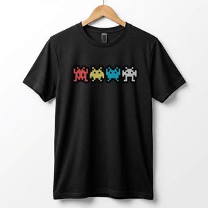 May include: Black t-shirt featuring a pixelated design of four colorful space invader-style aliens in red, yellow, blue, and white. The design is centered on the front of the shirt, which is hanging on a wooden hanger.