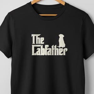 Op de afbeelding: Zwart T-shirt met de tekst "The Labfather" en een silhouet van een hond in vintage stijl. Het shirt hangt aan een houten hanger. Het ontwerp is in een distressed stijl, met een neutrale achtergrond.