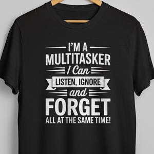 May include: Black t-shirt with the white text: "I'M A MULTITASKER I Can LISTEN, IGNORE and FORGET ALL AT THE SAME TIME!". The shirt is hanging on a wooden hanger.