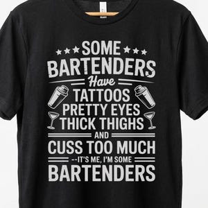 Puede incluir: Camiseta negra con texto blanco. El texto dice "SOME BARTENDERS Have TATTOOS PRETTY EYES THICK THIGHS AND CUSS TOO MUCH --IT'S ME, I'M SOME BARTENDERS". La camiseta está colgada en una percha de madera.