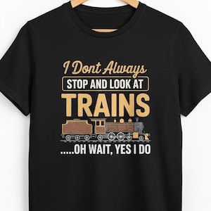 May include: Black t-shirt with the text "I Don't Always Stop and Look at Trains... Oh Wait, Yes I Do" in yellow and white. A brown and tan train graphic is below the text. The shirt is hanging on a wooden hanger.