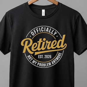 Könnte beinhalten: Schwarzes T-Shirt mit einem kreisförmigen Design, das "Officially Retired Est. 2026 Not My Problem Anymore" in goldfarbenen und weißen Buchstaben zeigt. Das Shirt hängt an einem Holzbügel vor grauem Hintergrund.
