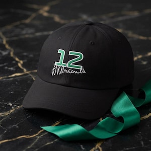 Peut inclure: Casquette de baseball noire avec une visière incurvée. Le chapeau porte le numéro "12" brodé en vert et blanc, ainsi qu'un texte stylisé. Un ruban vert est posé à côté.