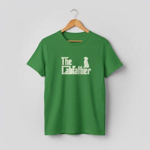 Op de afbeelding: Een groen t-shirt met de tekst "The Labfather" in een witte, gestileerde letter, met een silhouet van een hond. Het shirt hangt aan een houten hanger tegen een neutrale achtergrond.