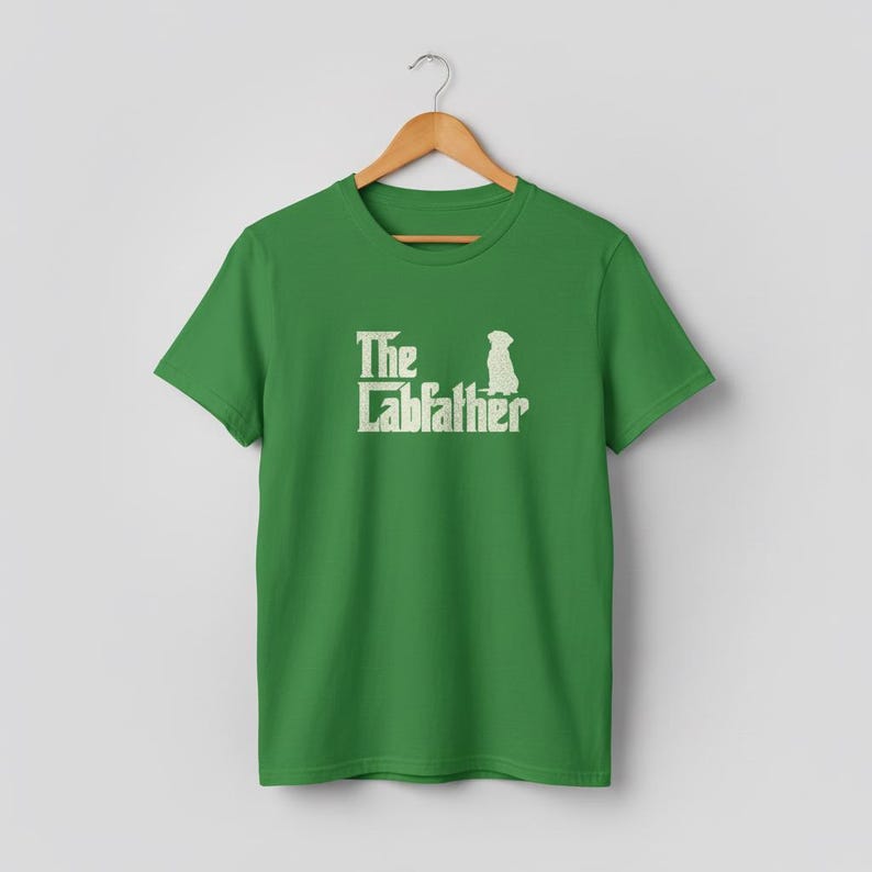 Op de afbeelding: Een groen t-shirt met de tekst "The Labfather" in een witte, gestileerde letter, met een silhouet van een hond. Het shirt hangt aan een houten hanger tegen een neutrale achtergrond.