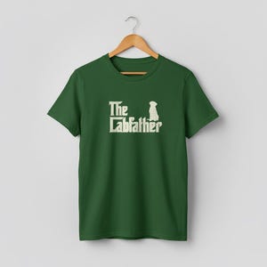Op de afbeelding: Groen t-shirt met de tekst "The Labfather" in een witte, verweerde lettertype, vergezeld van een silhouet van een hond. Het t-shirt hangt aan een houten hanger tegen een neutrale achtergrond. Het ontwerp is gecentreerd op de voorkant.