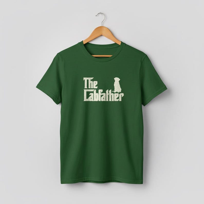Op de afbeelding: Groen t-shirt met de tekst "The Labfather" in een witte, verweerde lettertype, vergezeld van een silhouet van een hond. Het t-shirt hangt aan een houten hanger tegen een neutrale achtergrond. Het ontwerp is gecentreerd op de voorkant.