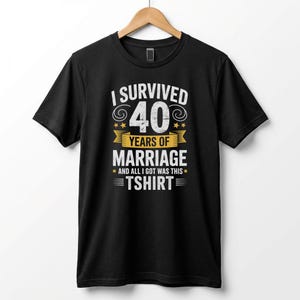 Op de afbeelding: Zwart T-shirt met de tekst "I Survived 40 Years of Marriage" in wit en geel. Het ontwerp bevat het getal 40 in een groot lettertype, met sterren en een banner. De tekst "And all I got was this T-shirt" is ook opgenomen.