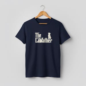 Op de afbeelding: Marineblauw T-shirt met de tekst "The Labfather" in een verweerde, gebroken witte lettertype. Een silhouet van een hond staat boven het woord "father". Het T-shirt hangt aan een houten hanger.