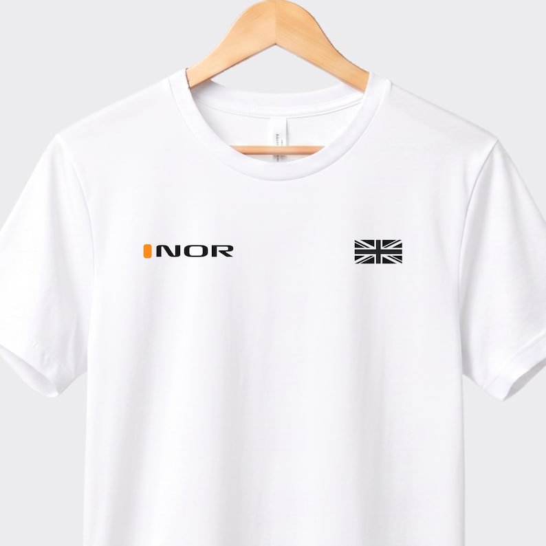 May include: White t-shirt featuring the word "INOR" in orange and black text on the left chest. A small black and white Union Jack flag is on the right chest. The shirt is displayed on a wooden hanger.