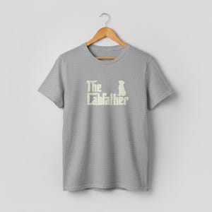 Op de afbeelding: Een lichtgrijs t-shirt met de tekst "The Labfather" in cr&egrave;me kleur, vergezeld van een silhouet van een hond. Het shirt hangt aan een houten hanger tegen een effen witte achtergrond.
