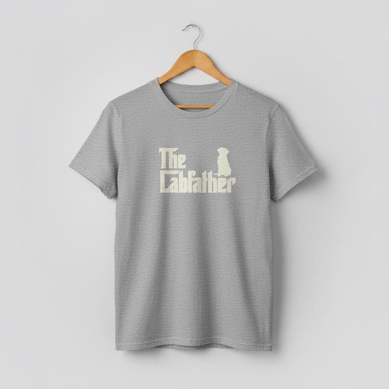 Op de afbeelding: Een lichtgrijs t-shirt met de tekst "The Labfather" in cr&egrave;me kleur, vergezeld van een silhouet van een hond. Het shirt hangt aan een houten hanger tegen een effen witte achtergrond.