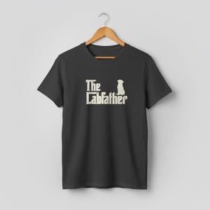 Op de afbeelding: Een zwart t-shirt met de tekst "The Labfather" in een witte, gestileerde lettertype, vergezeld van een silhouet van een hond. Het shirt hangt aan een houten hanger. Het ontwerp is een parodie op de film "The Godfather".