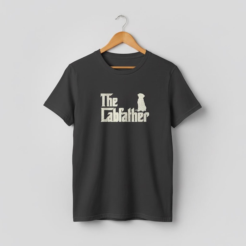 Op de afbeelding: Een zwart t-shirt met de tekst "The Labfather" in een witte, gestileerde lettertype, vergezeld van een silhouet van een hond. Het shirt hangt aan een houten hanger. Het ontwerp is een parodie op de film "The Godfather".