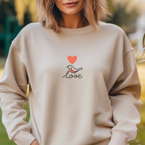 May include: A beige sweatshirt with a simple design of a red heart, a bird, and the word "love" in black.