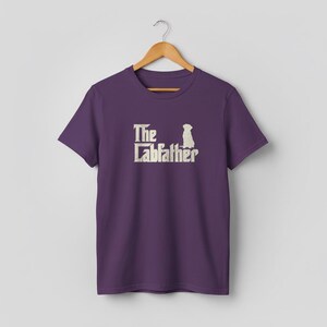Op de afbeelding: Paarse t-shirt met de tekst "The Labfather" en een honden silhouet in cr&egrave;me. Het t-shirt hangt aan een houten hanger tegen een neutrale achtergrond.