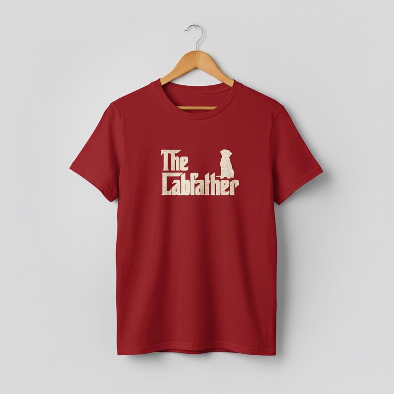 Op de afbeelding: Rood T-shirt met de tekst "The Labfather" en een honden silhouet in lichtbeige. Het T-shirt hangt aan een houten hanger. Het ontwerp is een parodie op een bekende filmtitel.