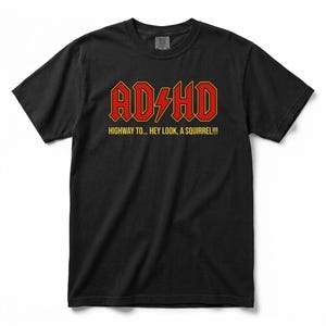 Könnte beinhalten: Ein schwarzes Kurzarm-T-Shirt mit dem roten Schriftzug ADHD und einem gelben Blitz in der Mitte. Unter dem Haupttext sind die gelben Worte HIGHWAY TO... HEY LOOK, A SQUIRREL!!! in einer serifenlosen Schrift auf der Brust aufgedruckt.