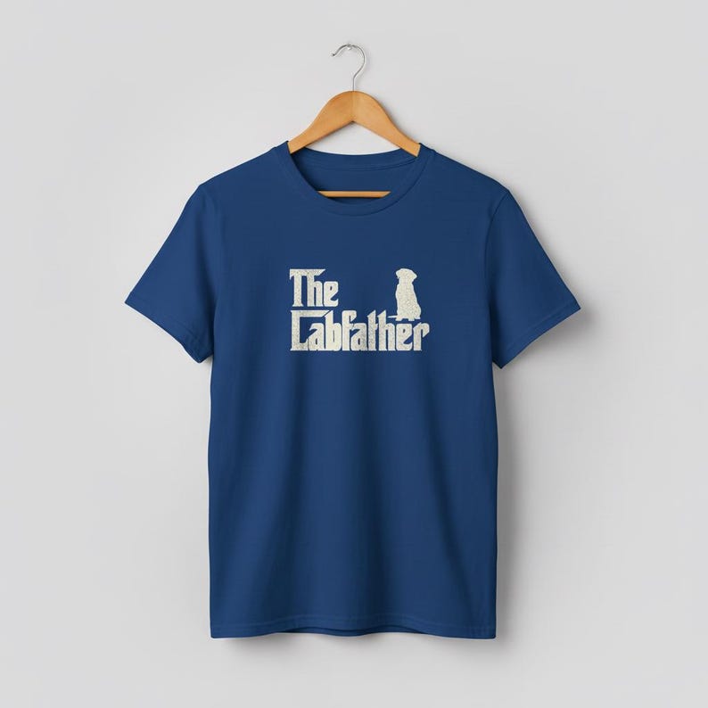 Op de afbeelding: Marineblauw T-shirt met de tekst "The Labfather" in verweerd gebroken wit en een silhouet van een hond. Het T-shirt hangt aan een houten kleerhanger.