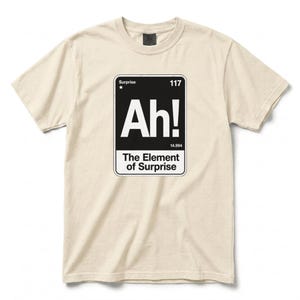 Op de afbeelding: Een crèmekleurig T-shirt met korte mouwen en een zwarte rechthoekige afbeelding die is ontworpen als een element uit het periodiek systeem. De afbeelding toont de tekst 'Surprise', '117', 'Ah!', '14.994' en 'The Element of Surprise' in witte letters.
