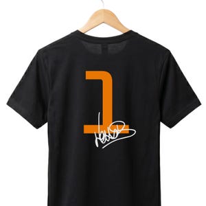 May include: Black t-shirt featuring an orange number 1 and a white signature. The garment is displayed on a wooden hanger. The design is on the back of the shirt.