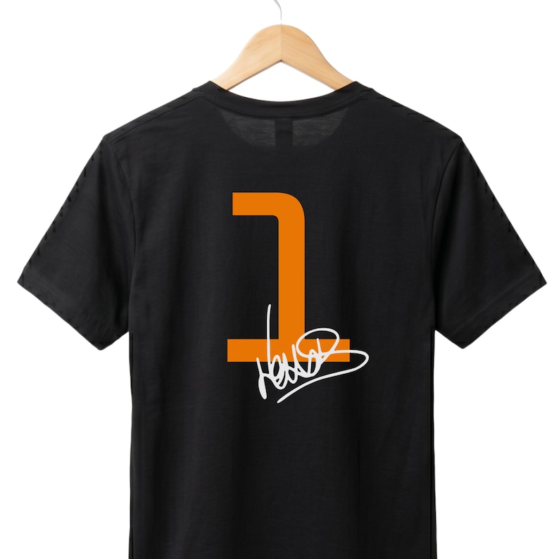 May include: Black t-shirt featuring an orange number 1 and a white signature. The garment is displayed on a wooden hanger. The design is on the back of the shirt.