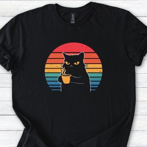 Może przedstawiać: Czarny t-shirt z grafiką zrzędliwego czarnego kota trzymającego filiżankę kawy. Projekt zawiera retro zachód słońca z czerwonymi, pomarańczowymi, żółtymi, niebieskimi i turkusowymi paskami. Koszulka wykonana jest z miękkiego materiału.