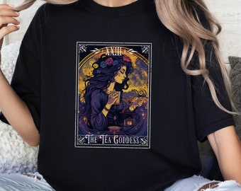 Puede incluir: Una camiseta negra con una ilustración colorida de una mujer con el pelo largo y oscuro, que lleva una corona de flores, sosteniendo una taza y una tetera. La ilustración está rodeada de un marco decorativo con el texto "The Tea Goddess" en la parte inferior.