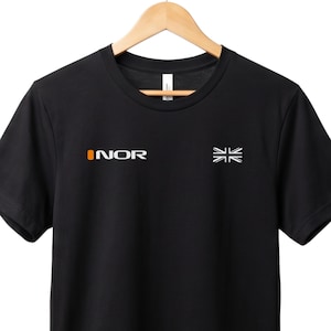 May include: Black t-shirt featuring the word "INOR" in white and orange on the left chest, and a white flag graphic on the right. The shirt is displayed on a wooden hanger.