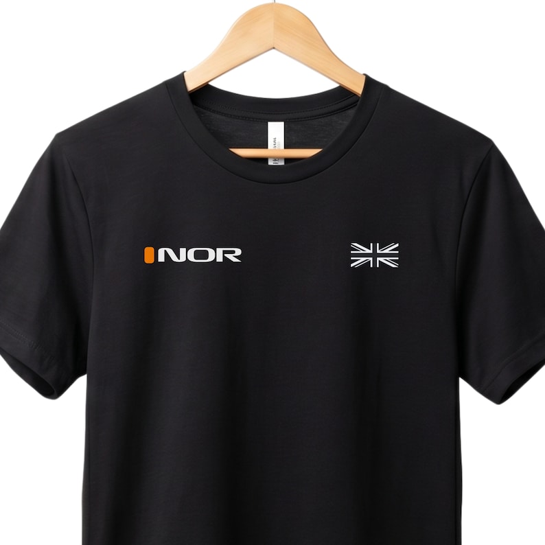 May include: Black t-shirt featuring the word "INOR" in white and orange on the left chest, and a white flag graphic on the right. The shirt is displayed on a wooden hanger.