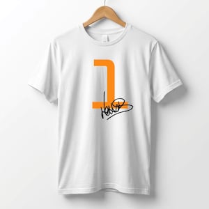 Puede incluir: Camiseta blanca con un diseño naranja del número 1 y una firma negra. El número 1 tiene una forma estilizada única. La camiseta está colgada en una percha de madera.