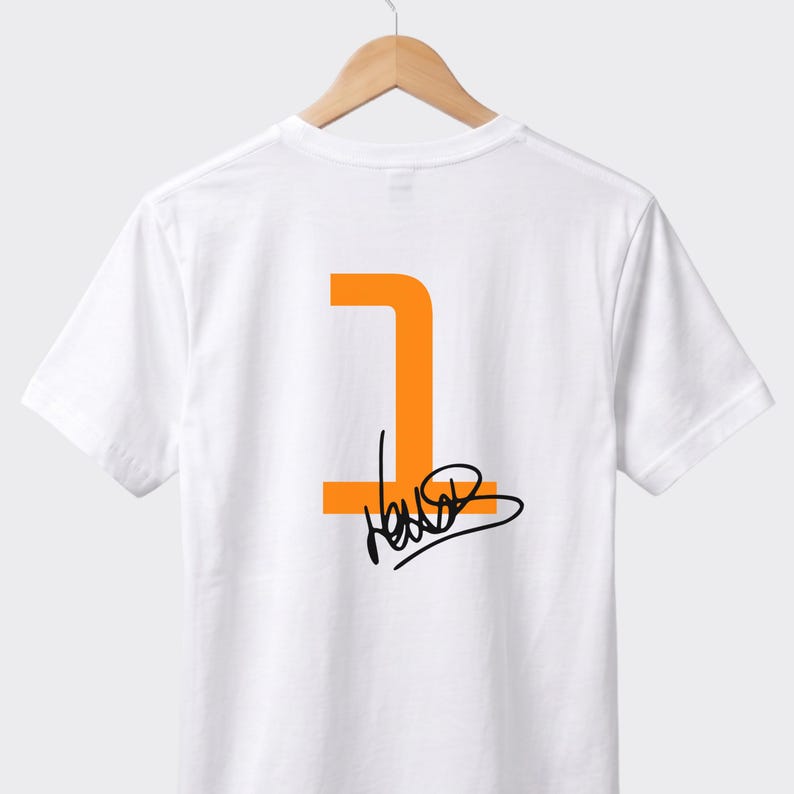 May include: White t-shirt featuring an orange number one and a black signature. The garment is suspended on a wooden hanger. The design is on the back of the shirt.